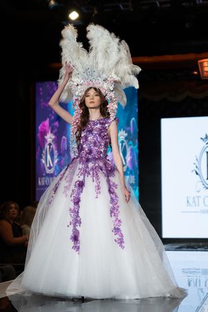 A luxurious white and purple ball gown with floral appliques and feather details glides down the fashion runway.