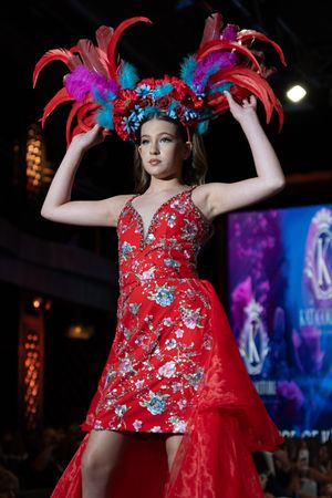 Model walks runway in red floral dress with feathered headpiece at fashion show under dramatic lighting.