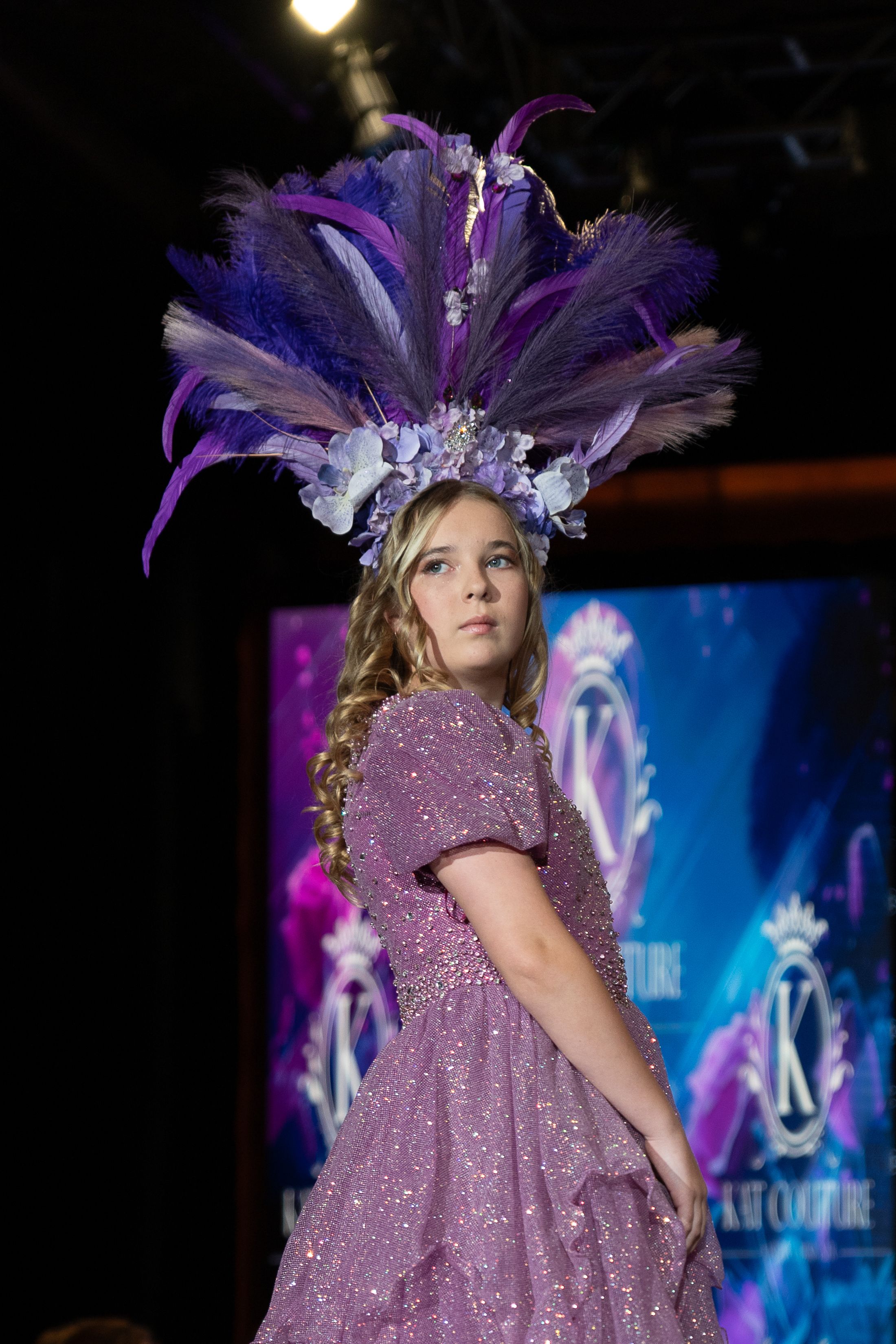 A model walks the runway wearing a sparkly purple dress and feathered headpiece against a blue-lit backdrop.