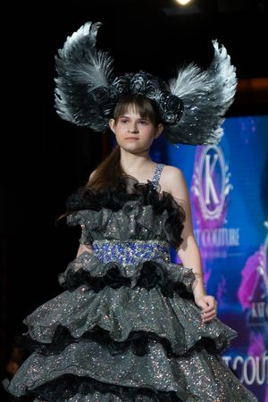 A fashion model walks the runway in a sparkly black tulle dress with dramatic feathered headpiece at a couture show.