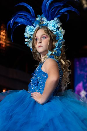 A model in a sparkling blue ball gown with feathered headpiece and floral crown poses during a fashion show.