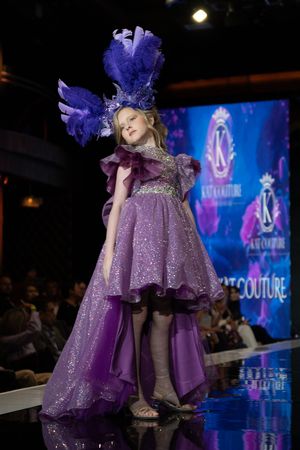 A model walks the runway in a purple high-low gown with matching feathered headpiece at a couture fashion show.