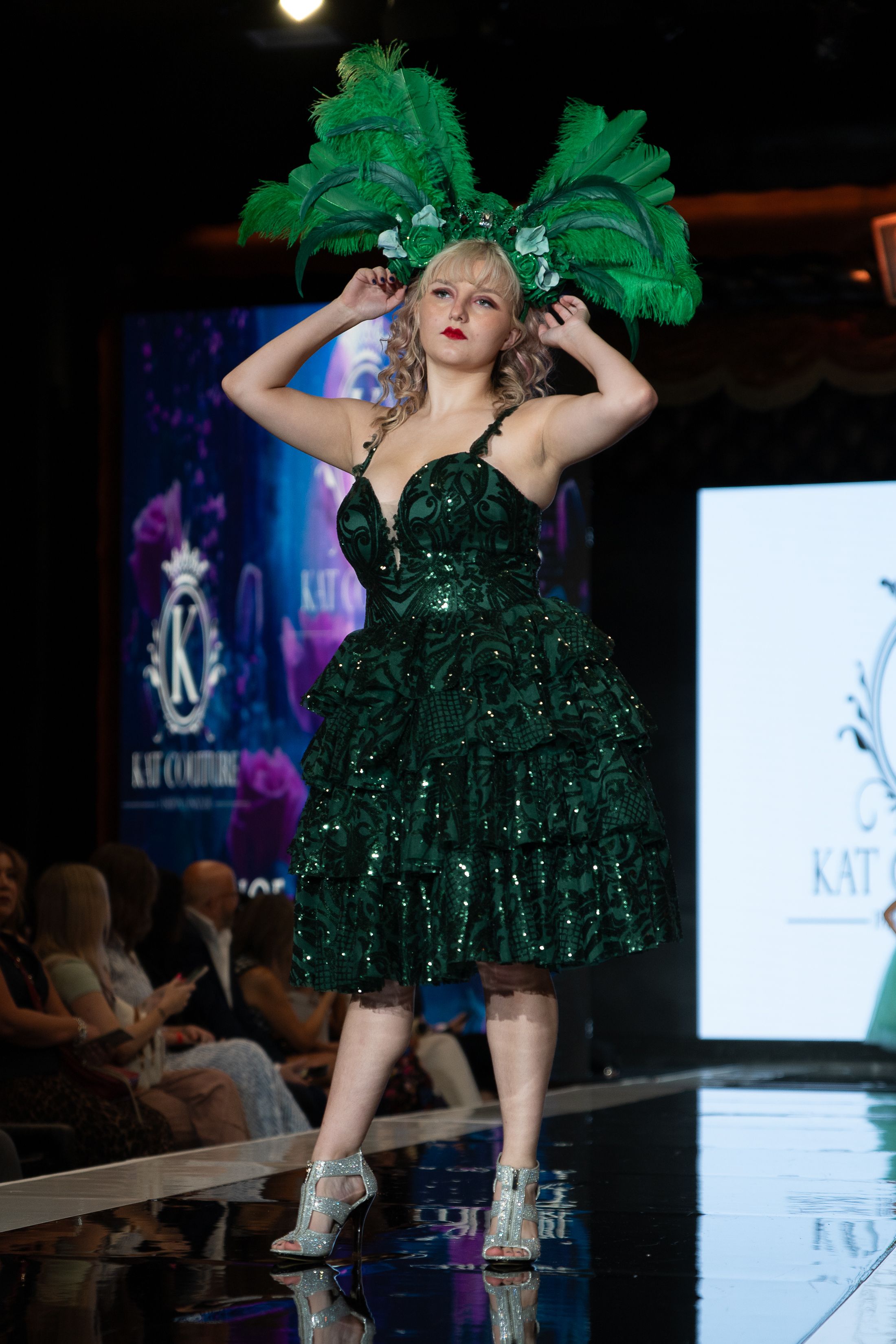 Model struts down runway in sparkling emerald green cocktail dress with feather details at fashion show.