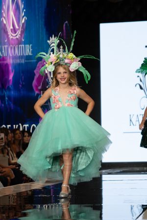 Model walks runway in mint green tulle dress with floral headpiece at fashion show event.