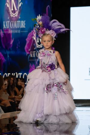 Young model walks runway in lavender tulle ball gown with purple floral embellishments at fashion show.