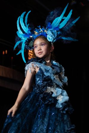 A performer in a dramatic blue feathered costume and headdress stands against a dark background.