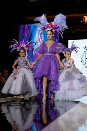Models showcase elegant white tulle and purple gowns with feathered headpieces on a dramatic runway stage.