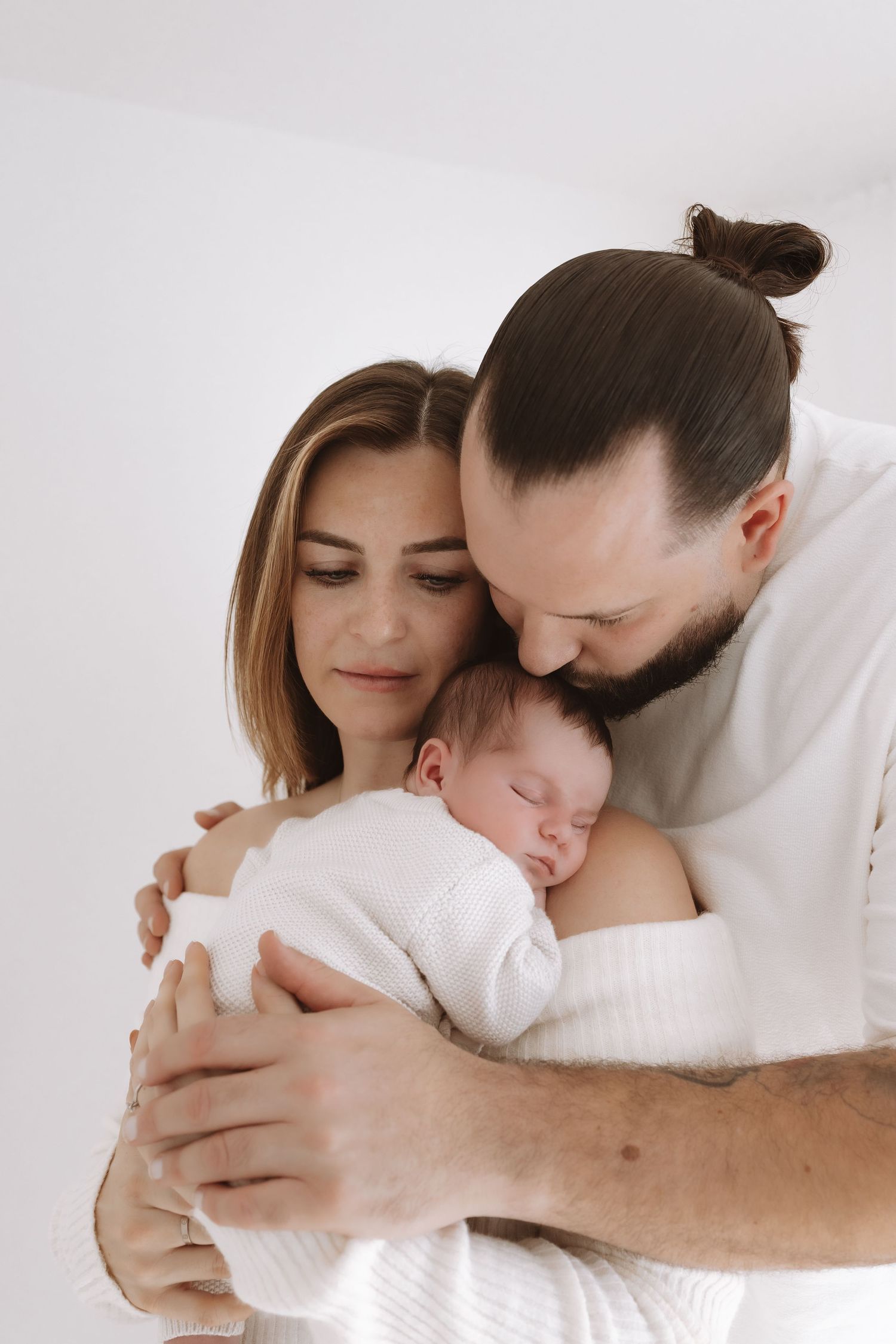 Parents in white clothing tenderly embrace their sleeping newborn baby in an intimate family portrait.
