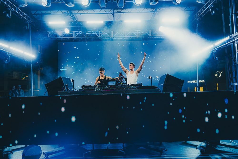 DJ performs on stage at an electronic music festival with dramatic blue lighting and smoke effects.