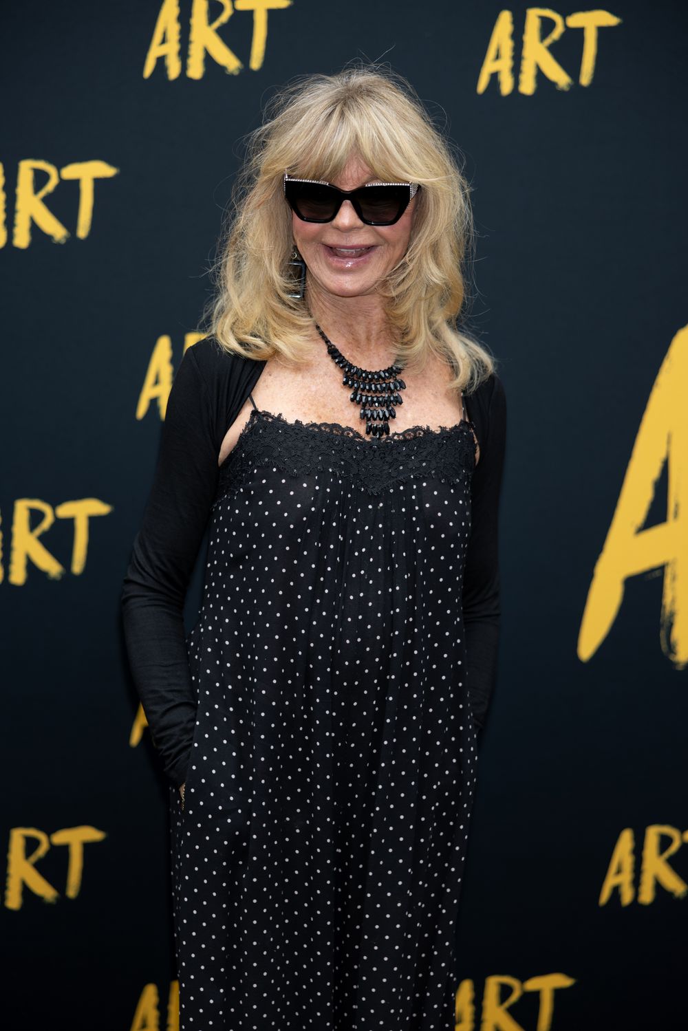Person in polka dot dress and sunglasses poses against dark backdrop with gold 'ART' text.