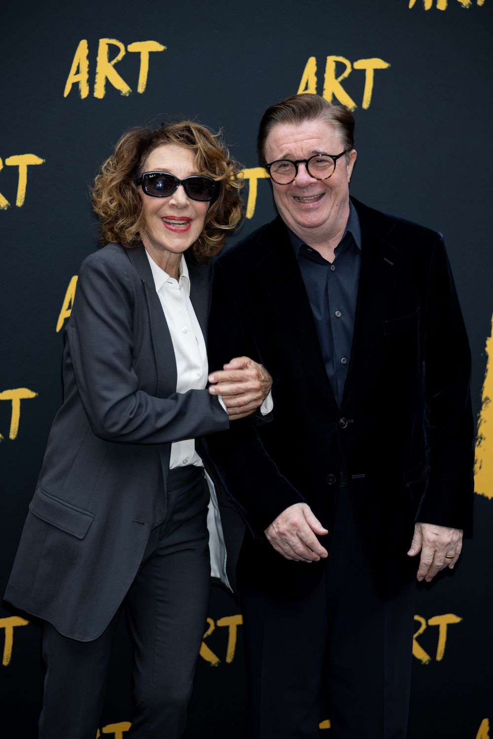Two people in formal black attire share a lighthearted moment against a dark backdrop with gold text.