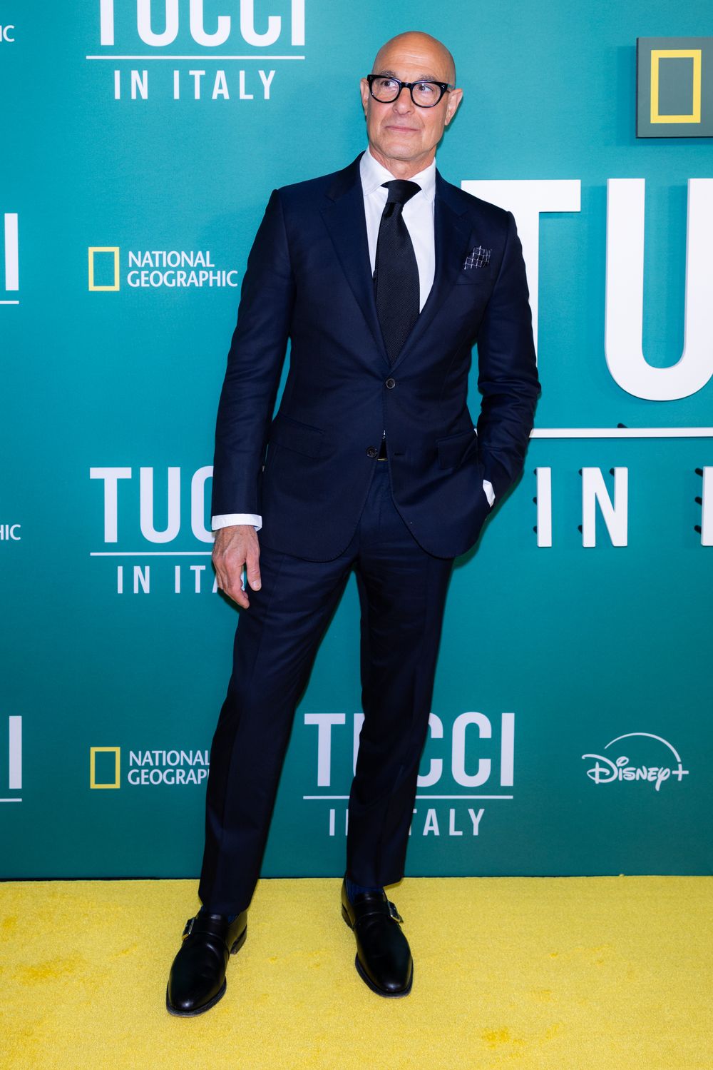 Person in a dark suit stands against a turquoise and yellow backdrop with National Geographic branding.