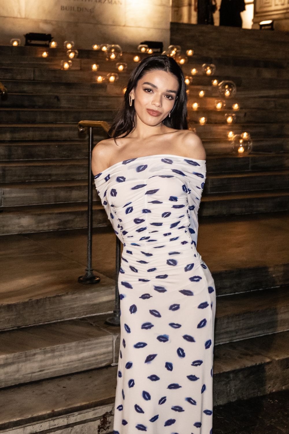 A person poses in an off-shoulder white dress with navy blue lip print pattern against a rustic wooden wall with string lights.
