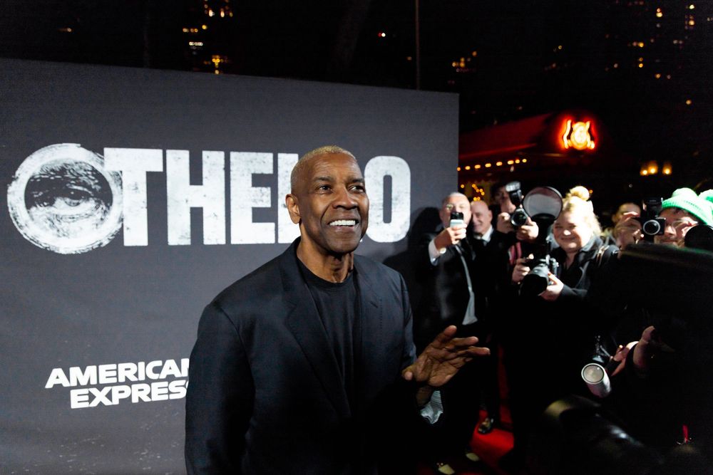 A smiling individual in a black sweater poses at a movie premiere event for 'Othello' with American Express branding visible.