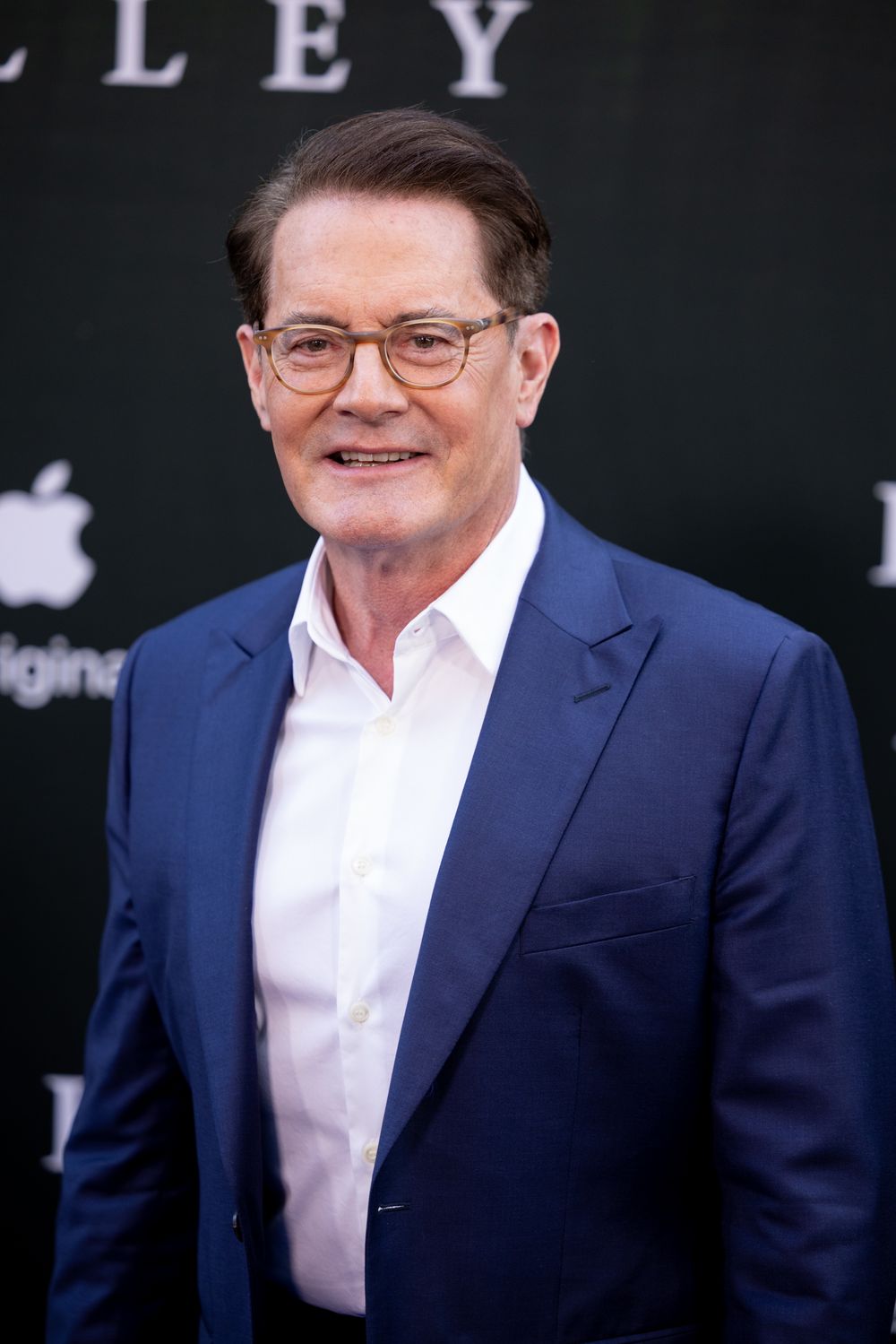 Person in navy suit and glasses poses at formal event against dark backdrop.