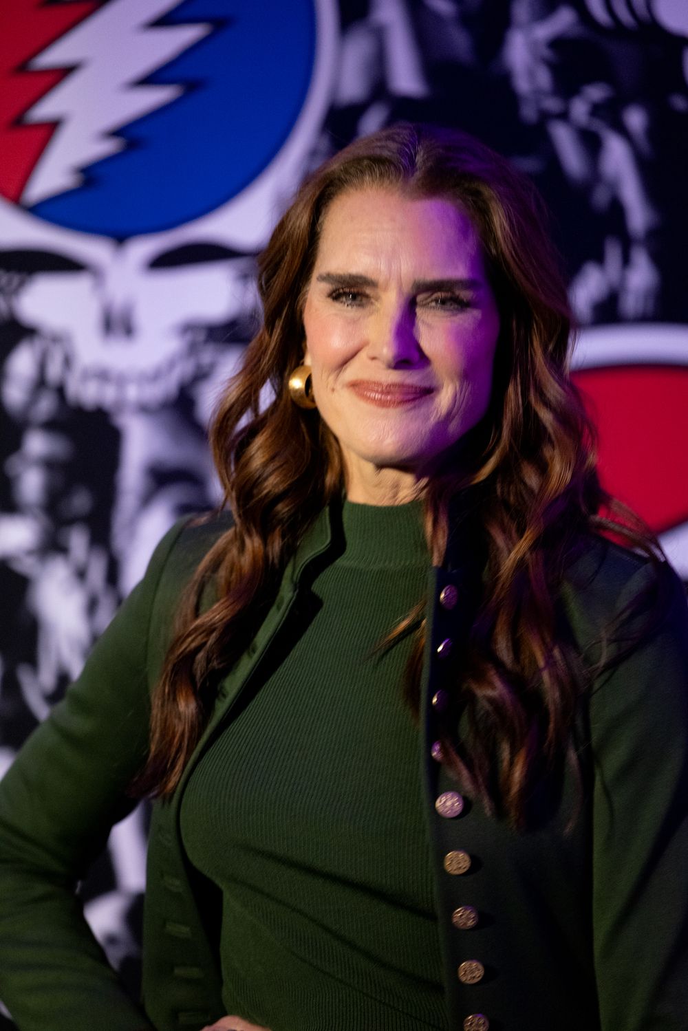A person wearing a dark green turtleneck sweater with buttons smiles while posing against a red and blue backdrop.