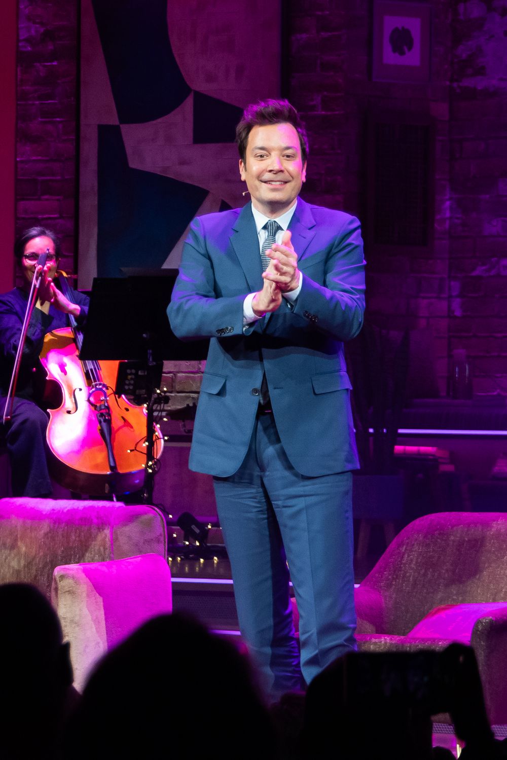 Performer in blue suit gestures on stage during nighttime concert with purple lighting and audience silhouettes visible.