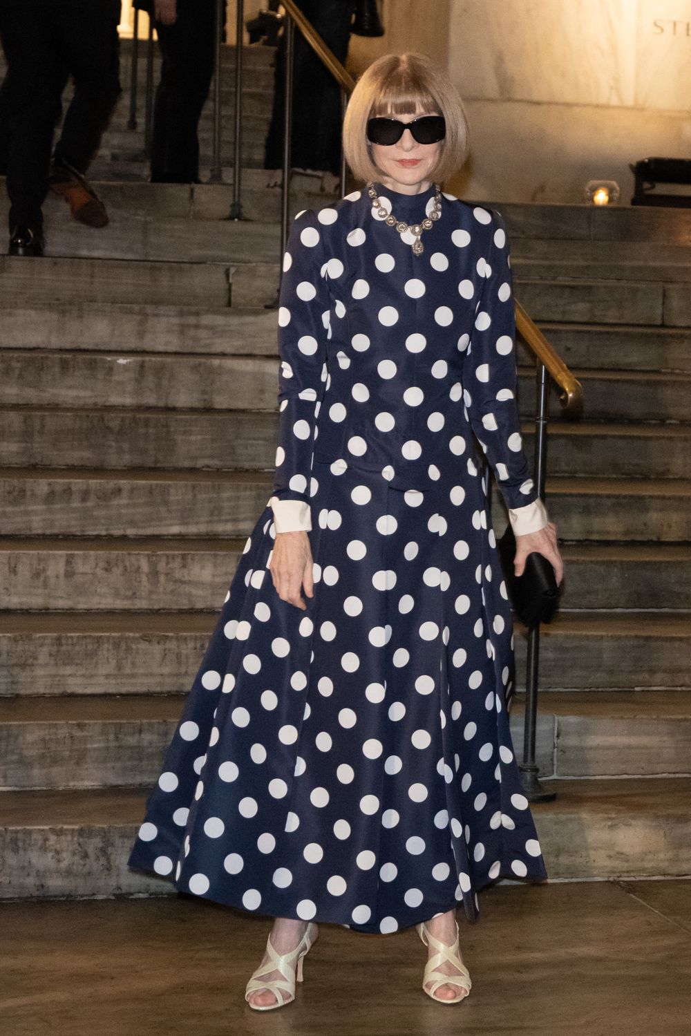 A navy blue and white polka dot dress with flared sleeves is shown from multiple angles on a marble staircase.