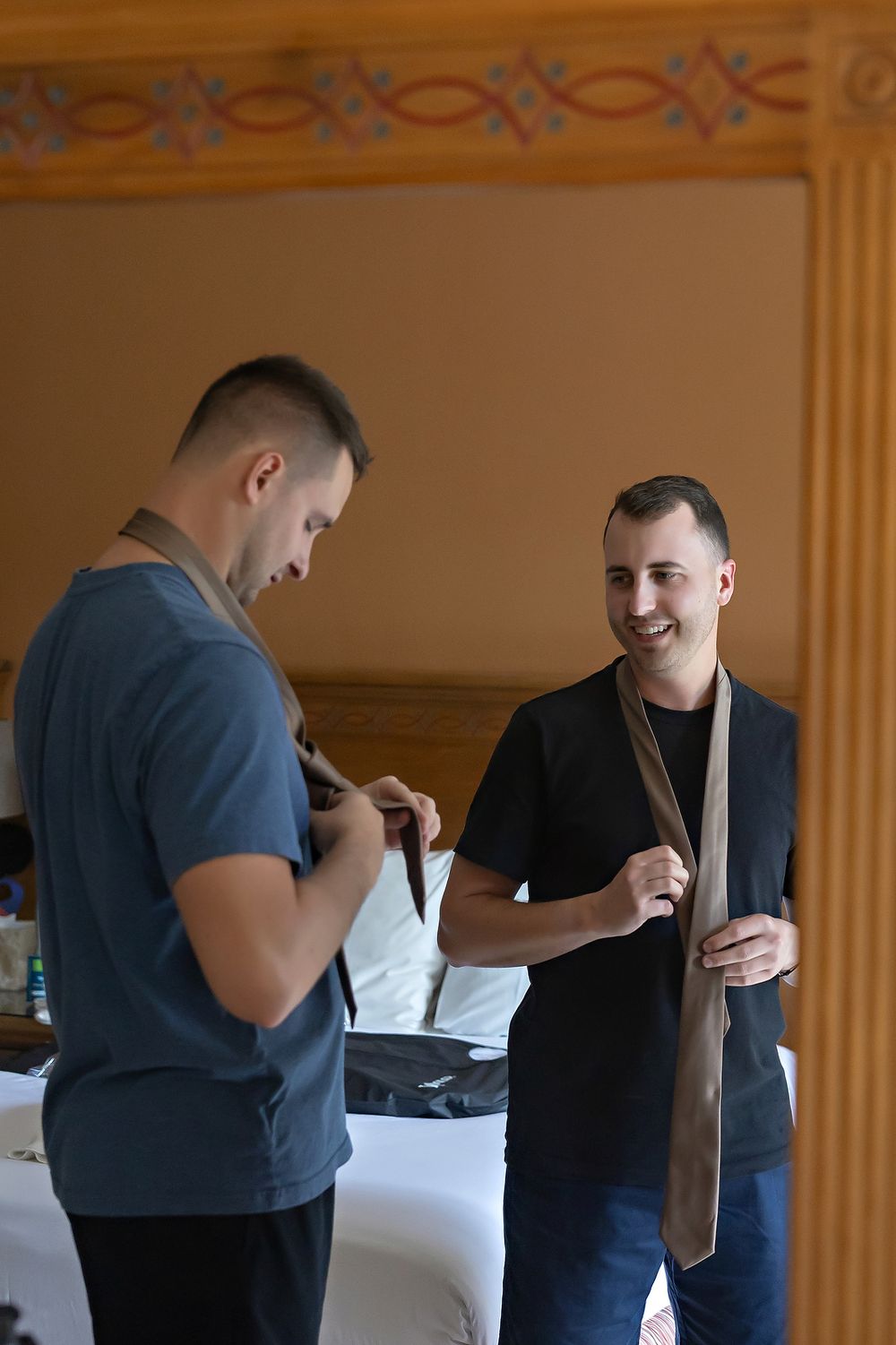 Two men stand in a room as one adjusts his tie while the other smiles during what appears to be preparation time.