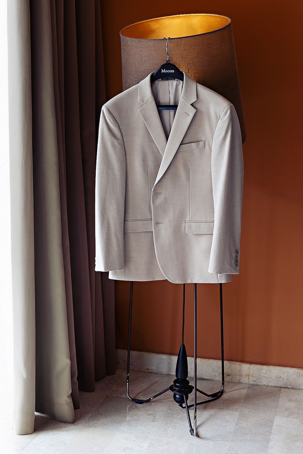 A light gray formal suit jacket displayed on a black clothing stand against a brown wall.