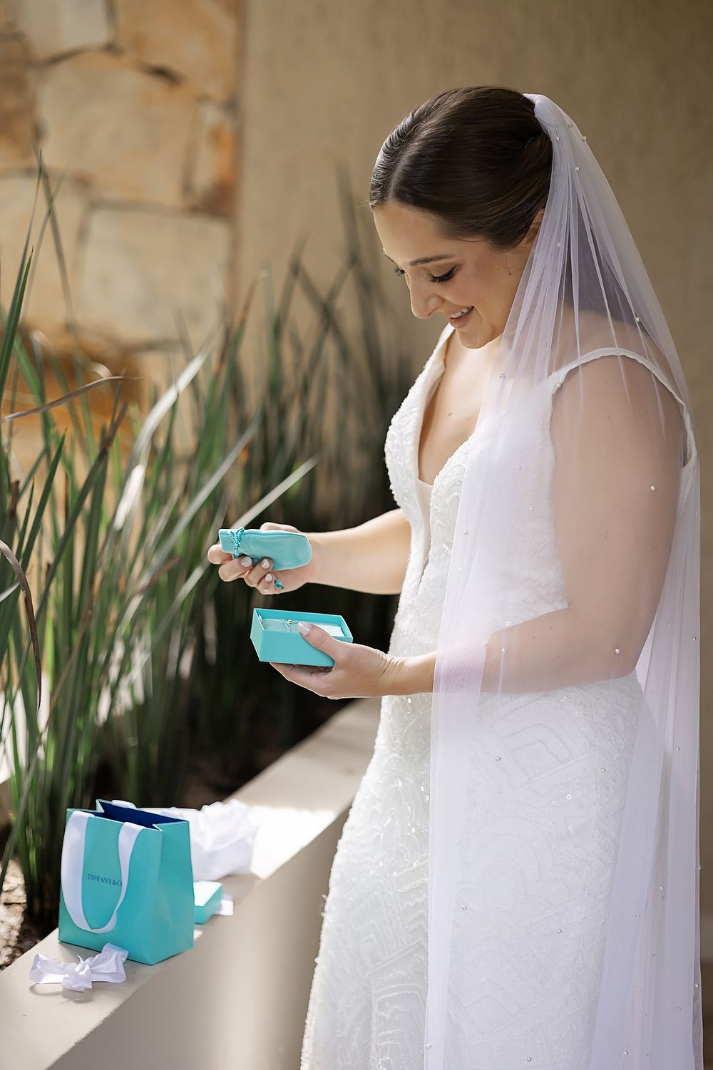 A bride in a white wedding dress and veil opens a small turquoise gift box in an outdoor garden setting.