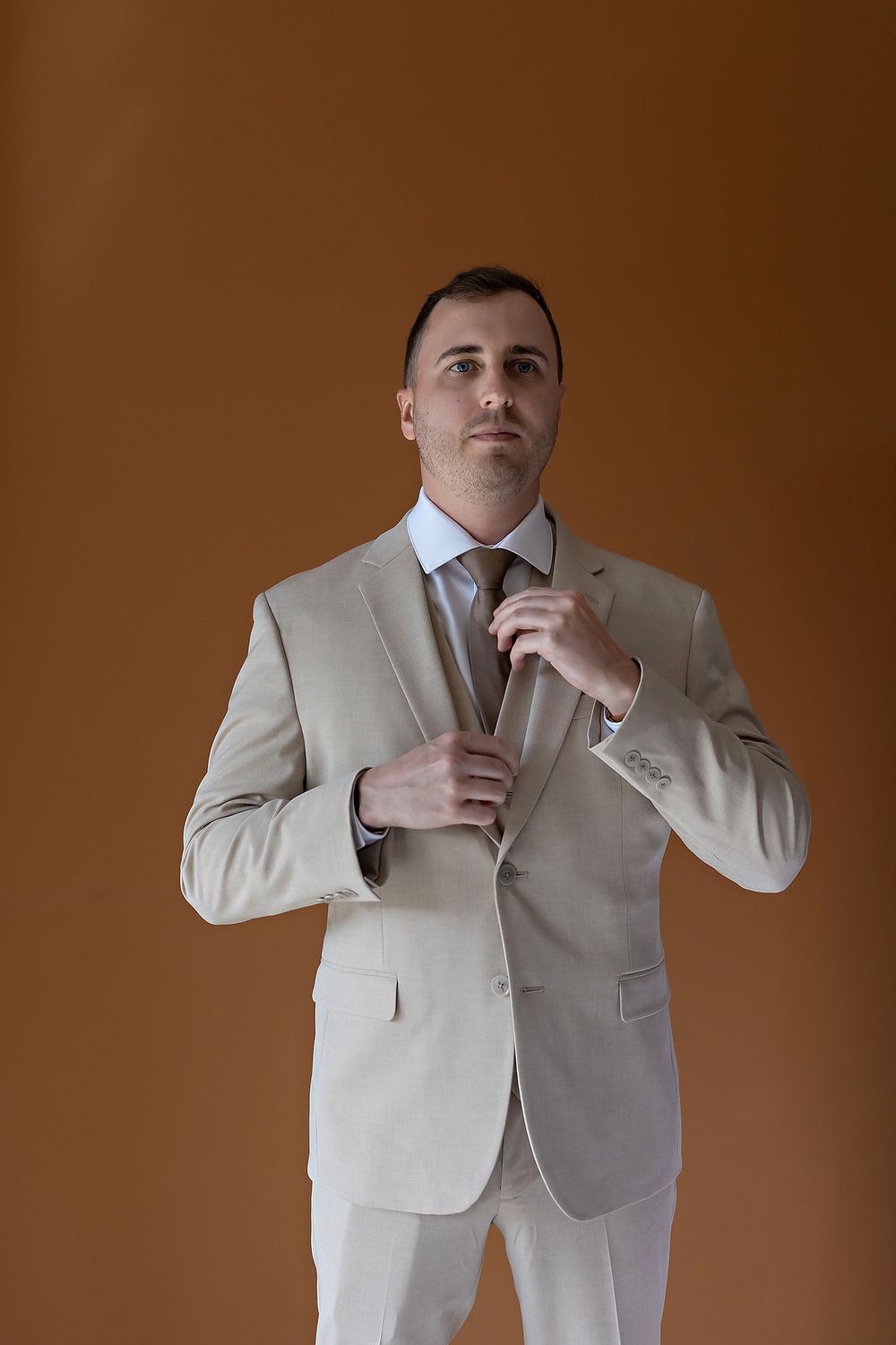 Professional wearing a light beige suit adjusts their tie and collar in a sequence of poses against a brown backdrop.