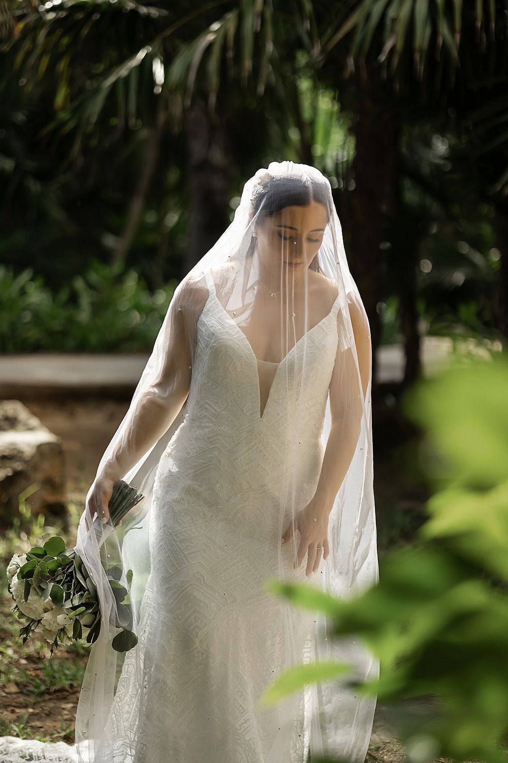 A bride in an elegant white lace wedding dress stands in a garden setting with soft natural lighting and greenery.