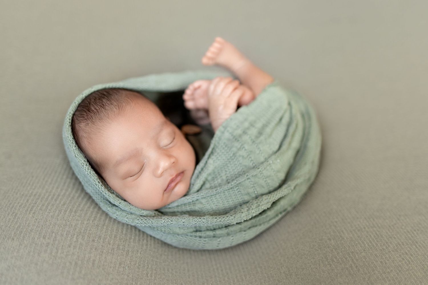 A sleeping newborn baby wrapped in a soft sage green swaddle blanket lies on a neutral beige backdrop.