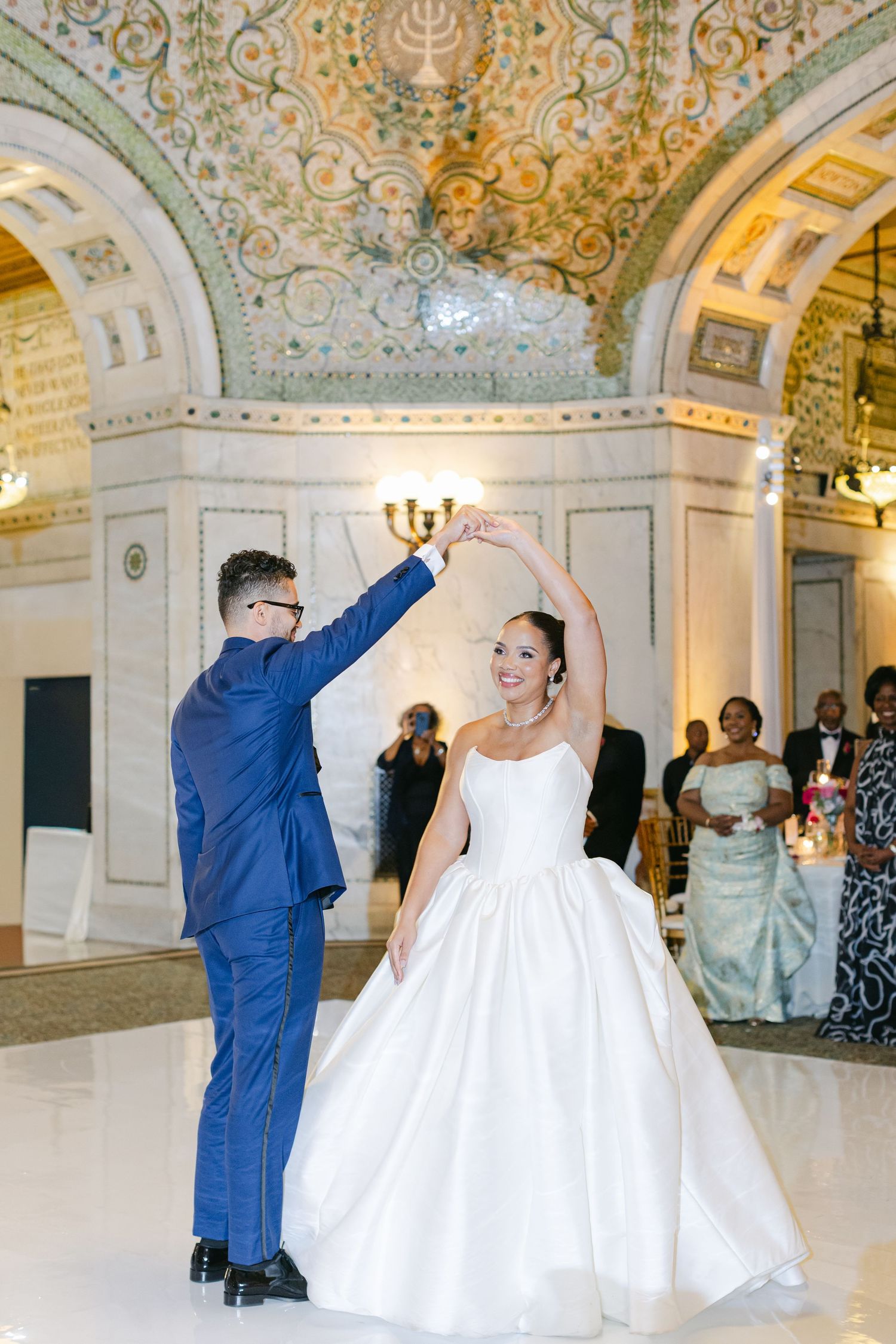 Dancing couples celebrate in an ornate ballroom with elegant architecture and decorative ceiling details.