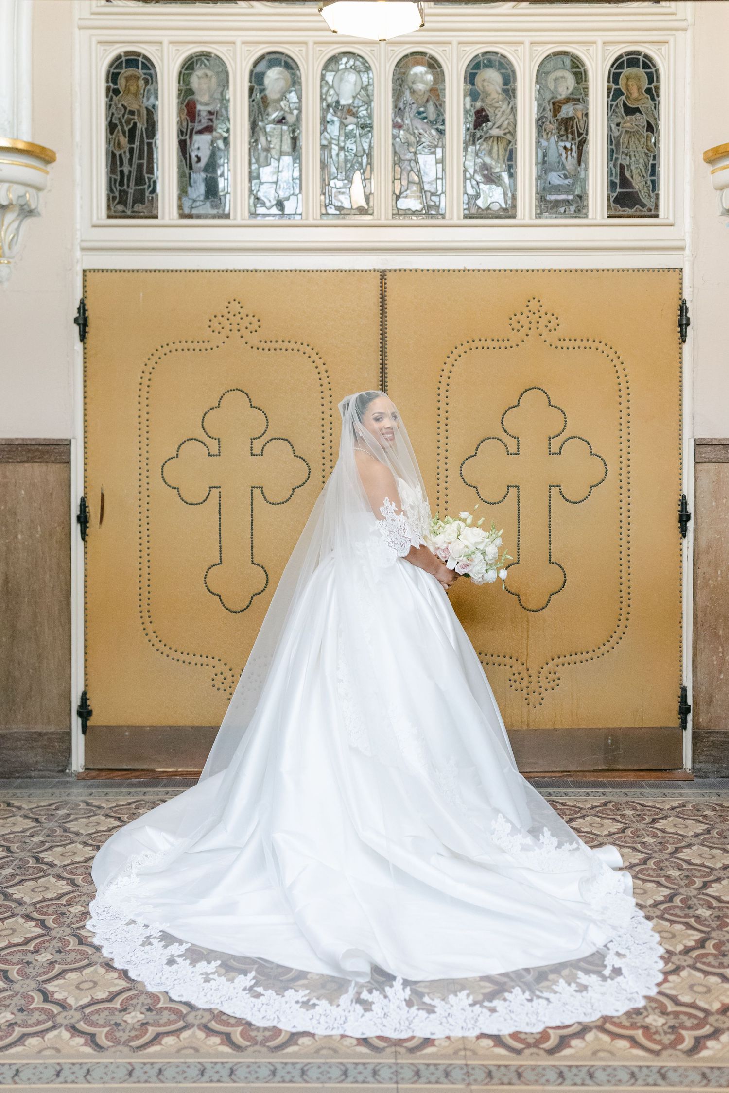 A bride in a flowing white ballgown and veil stands before ornate church doors with stained glass windows above.