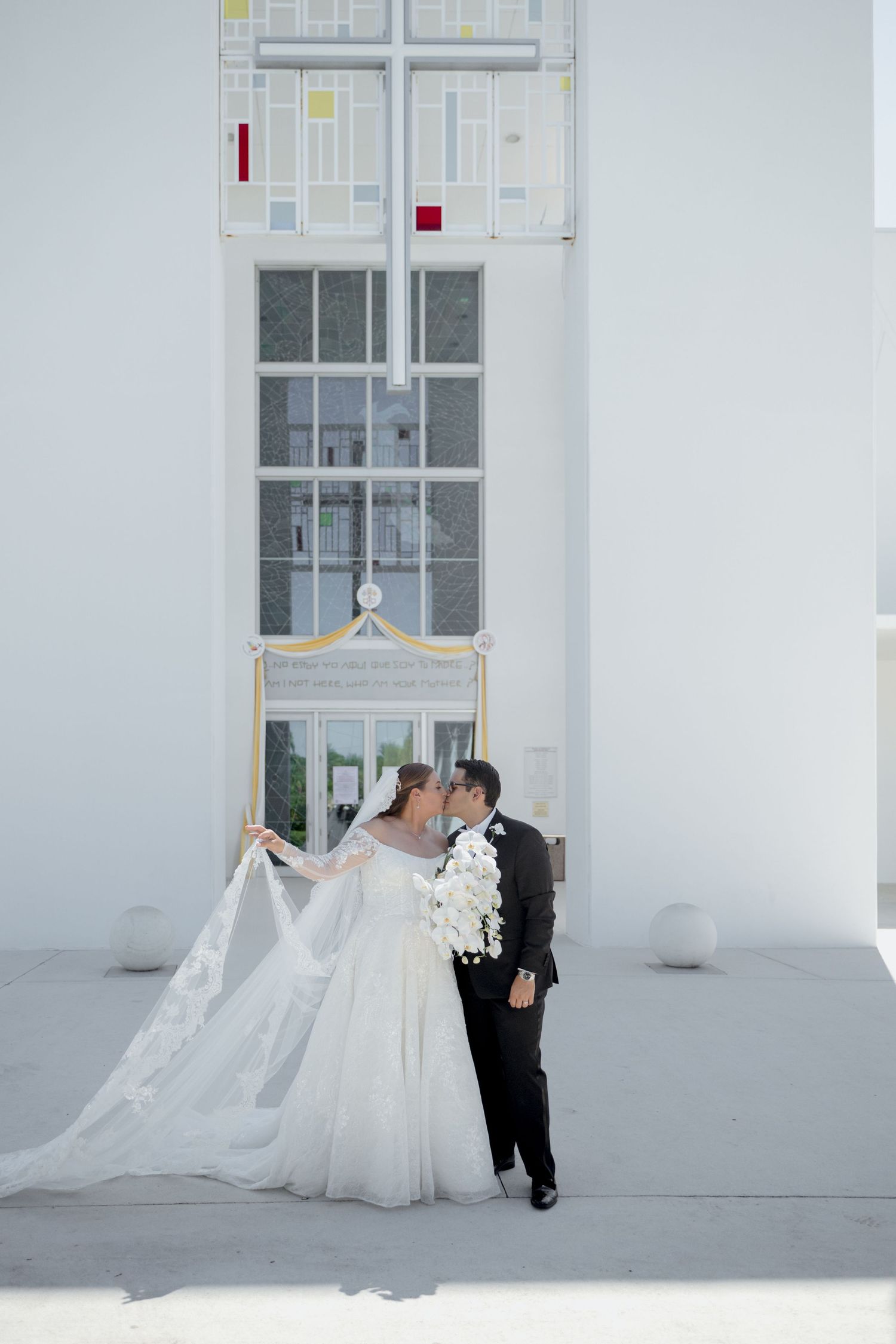 A couple in wedding attire share a romantic moment against a white building with tall windows and elegant architecture.