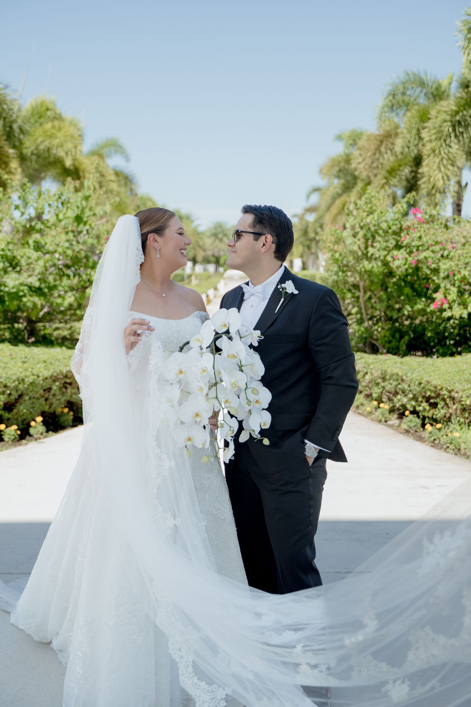 A romantic wedding couple poses outdoors with a flowing white veil and orchid bouquet against lush greenery.