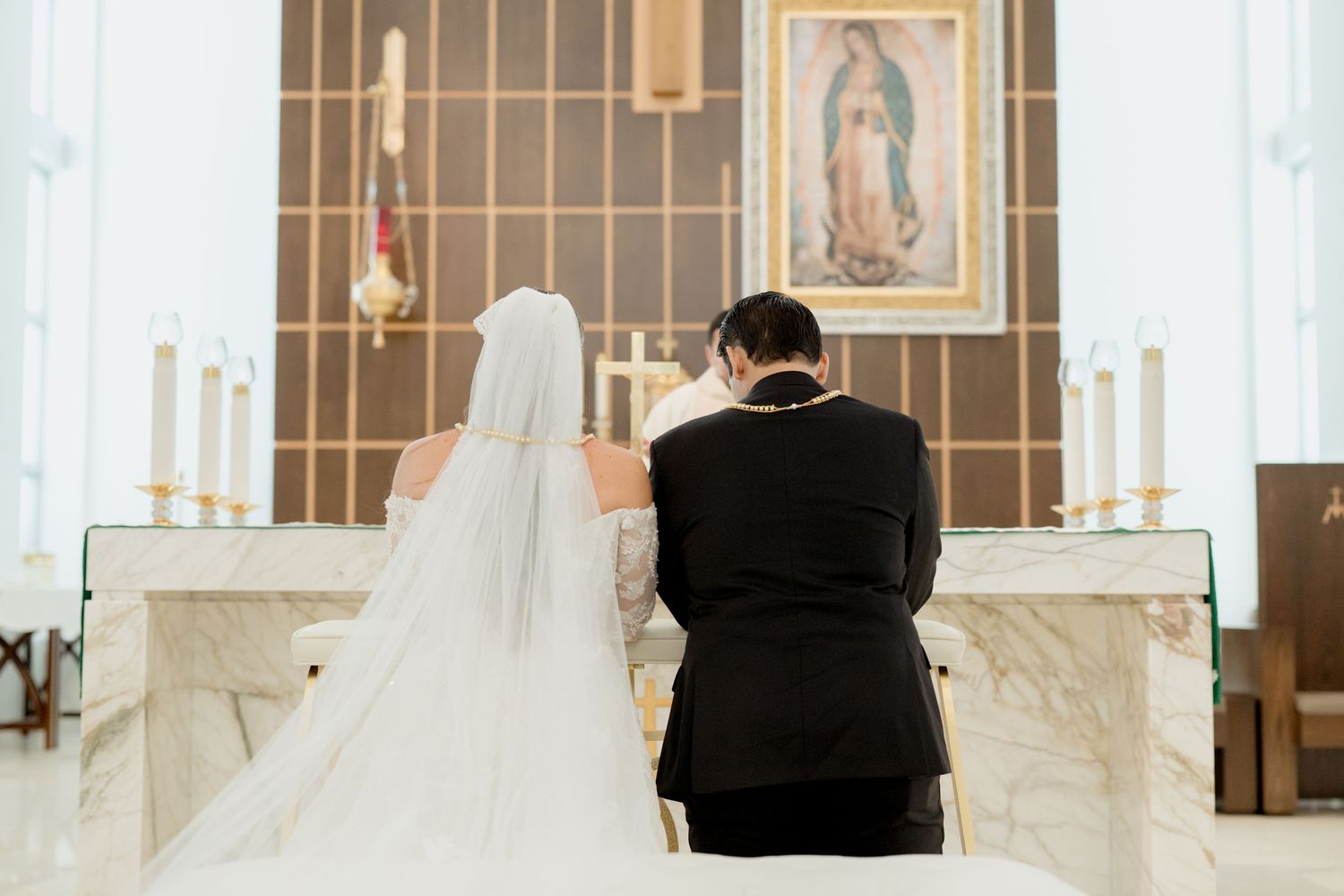 A couple kneels at a church altar during a wedding ceremony with a Virgin Mary painting in the background.