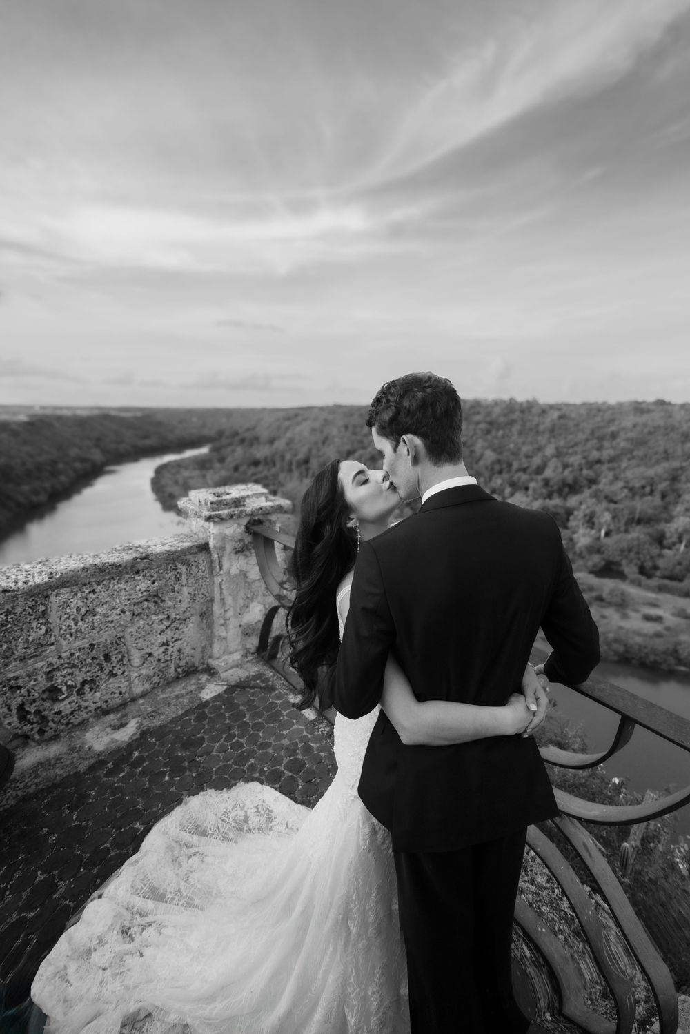 A romantic couple shares an intimate moment overlooking a scenic river valley in black and white photography.