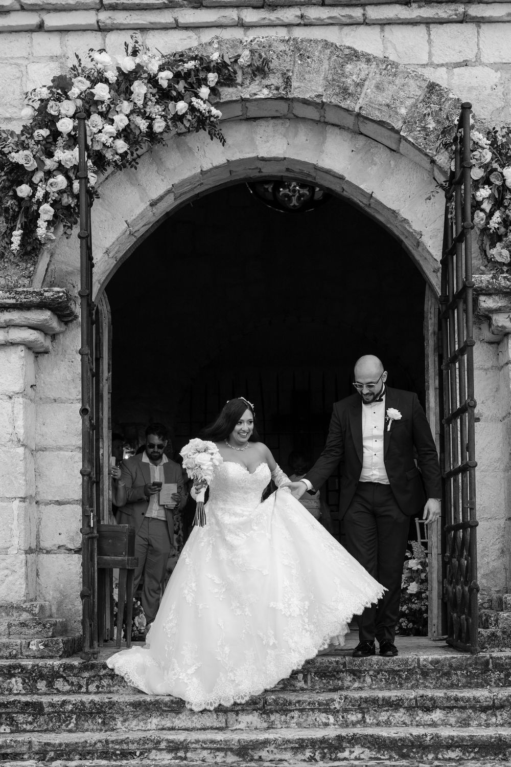 A sequence of black and white wedding photos showing a couple posing under stone archways decorated with flowers.