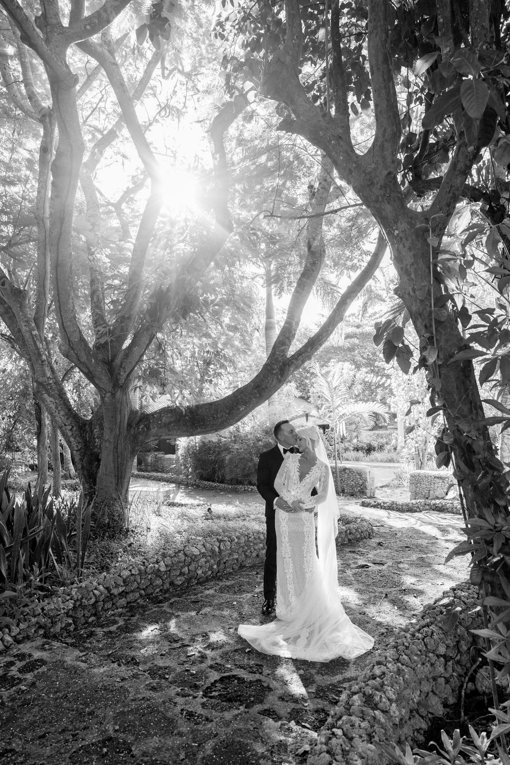 A romantic garden wedding scene captured in black and white with sunlight streaming through trees on a stone pathway.