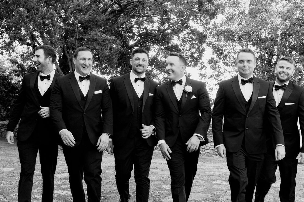 Wedding party members in black tuxedos walk together outdoors while laughing and smiling.