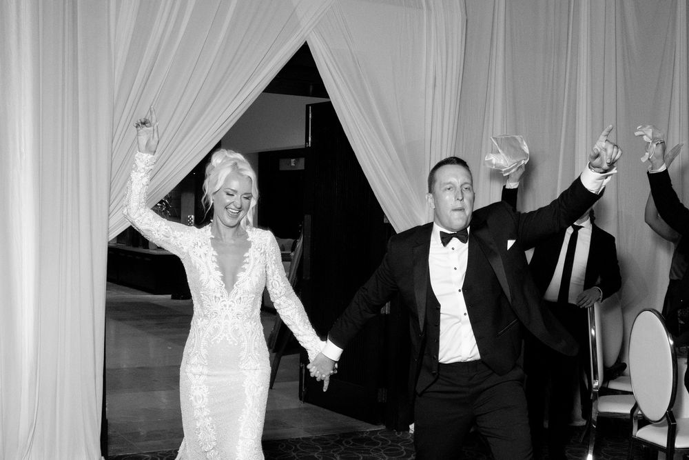 Black and white panoramic photo of people dancing with raised arms at a formal celebration event.