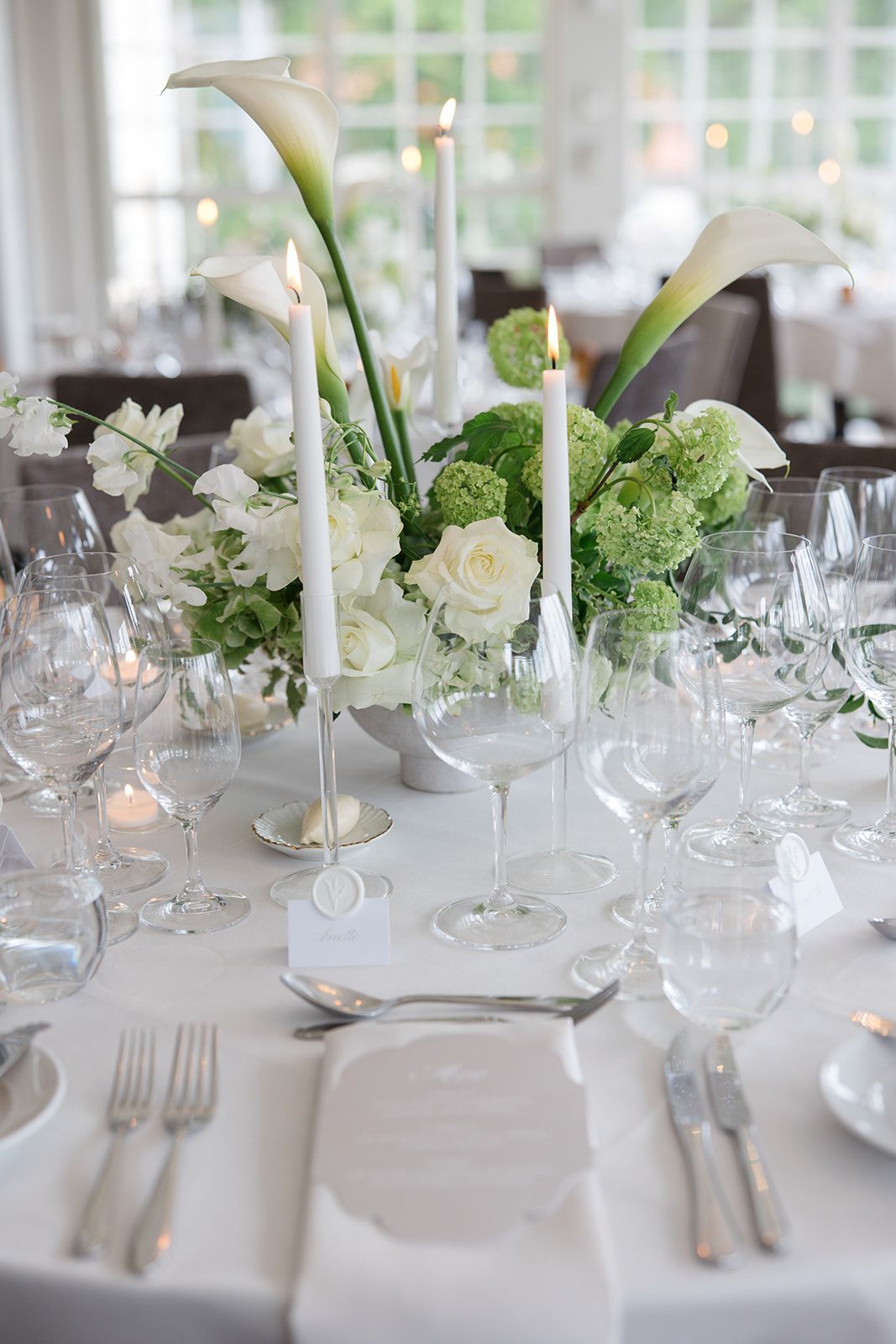 Elegant white wedding reception table settings with crystal glassware and delicate floral centerpieces in natural light.