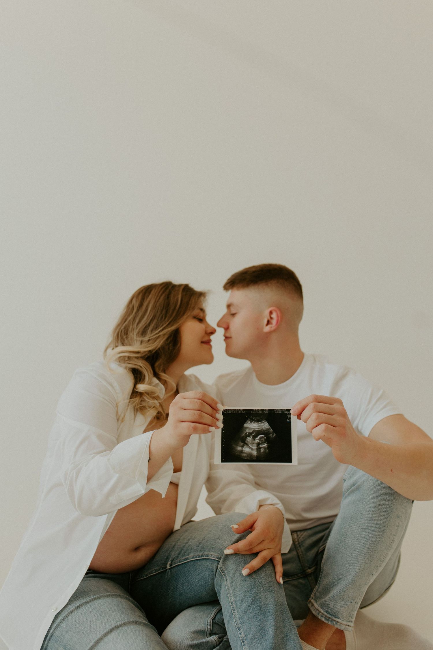 A couple in white and denim sits on a bed holding an ultrasound photo while sharing an intimate moment together.