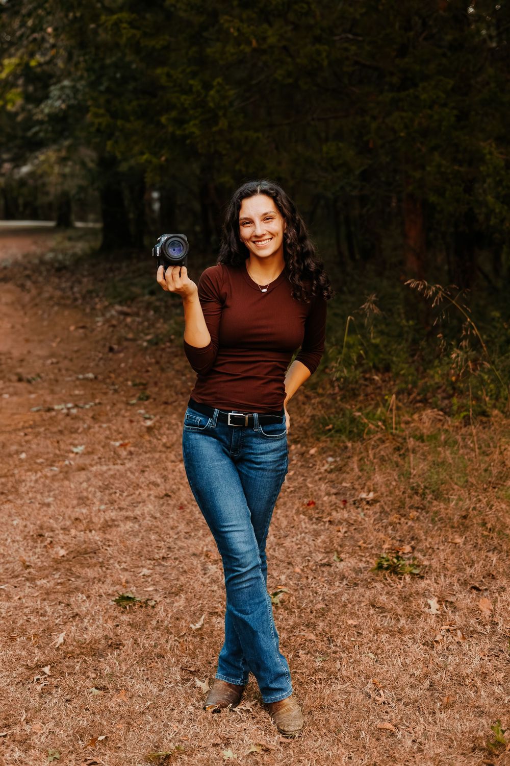 A photographer in jeans and burgundy top poses with camera equipment outdoors.