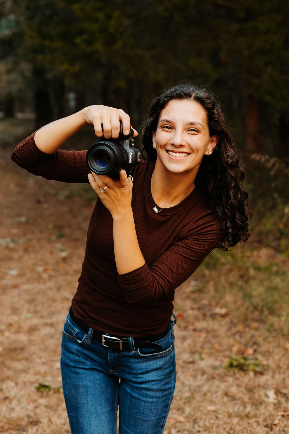 Photographer in brown top and jeans demonstrating creative camera angles outdoors.