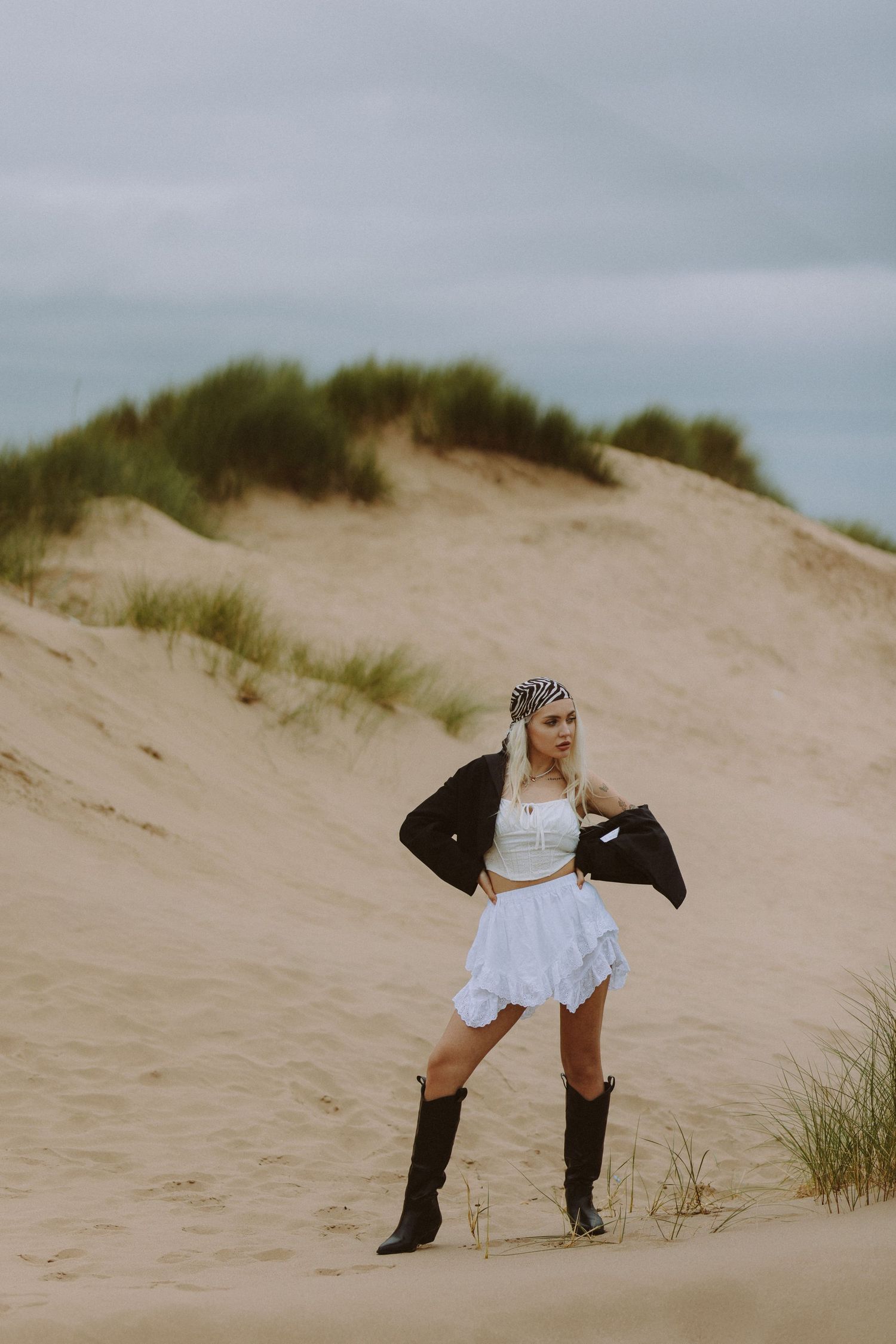 A person in a white dress and black boots poses artistically on sandy dunes with beach grass in the background.