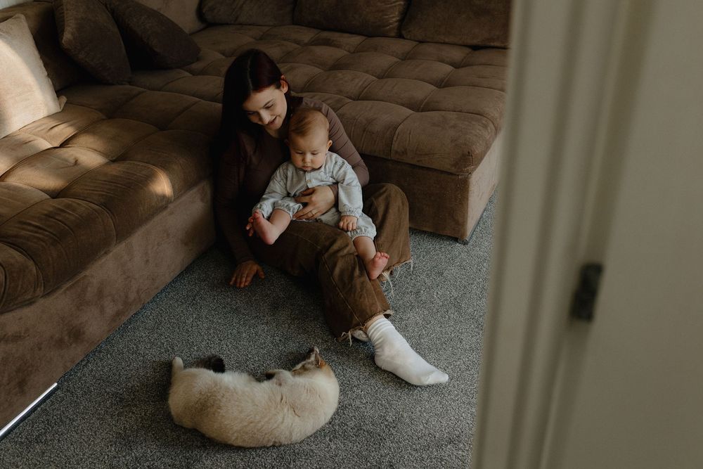 A parent and child sit together on the floor by a brown couch with a sleeping dog nearby in warm lighting.