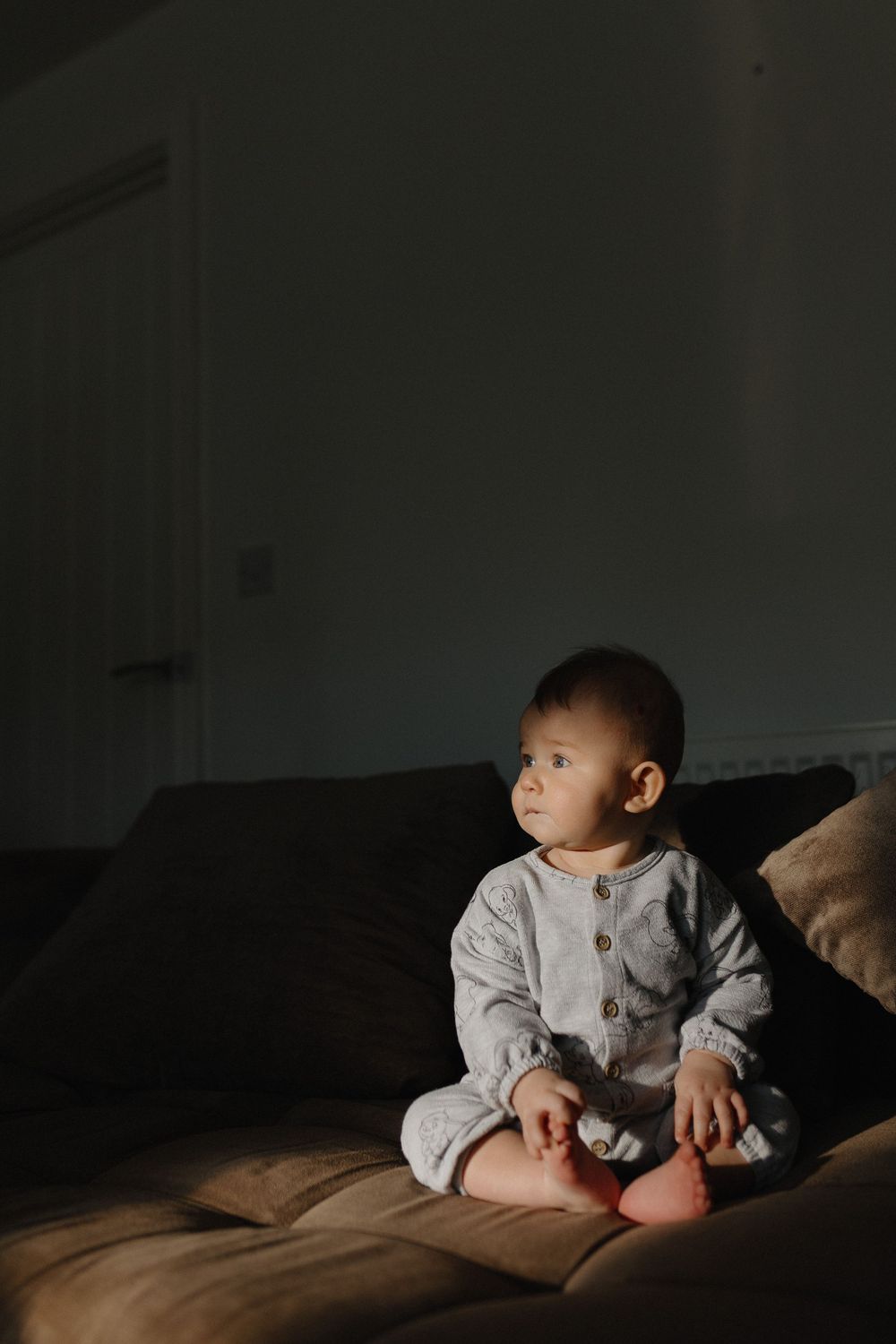 A baby in gray clothing lies on dark bedding while exploring and reaching in moody lighting.