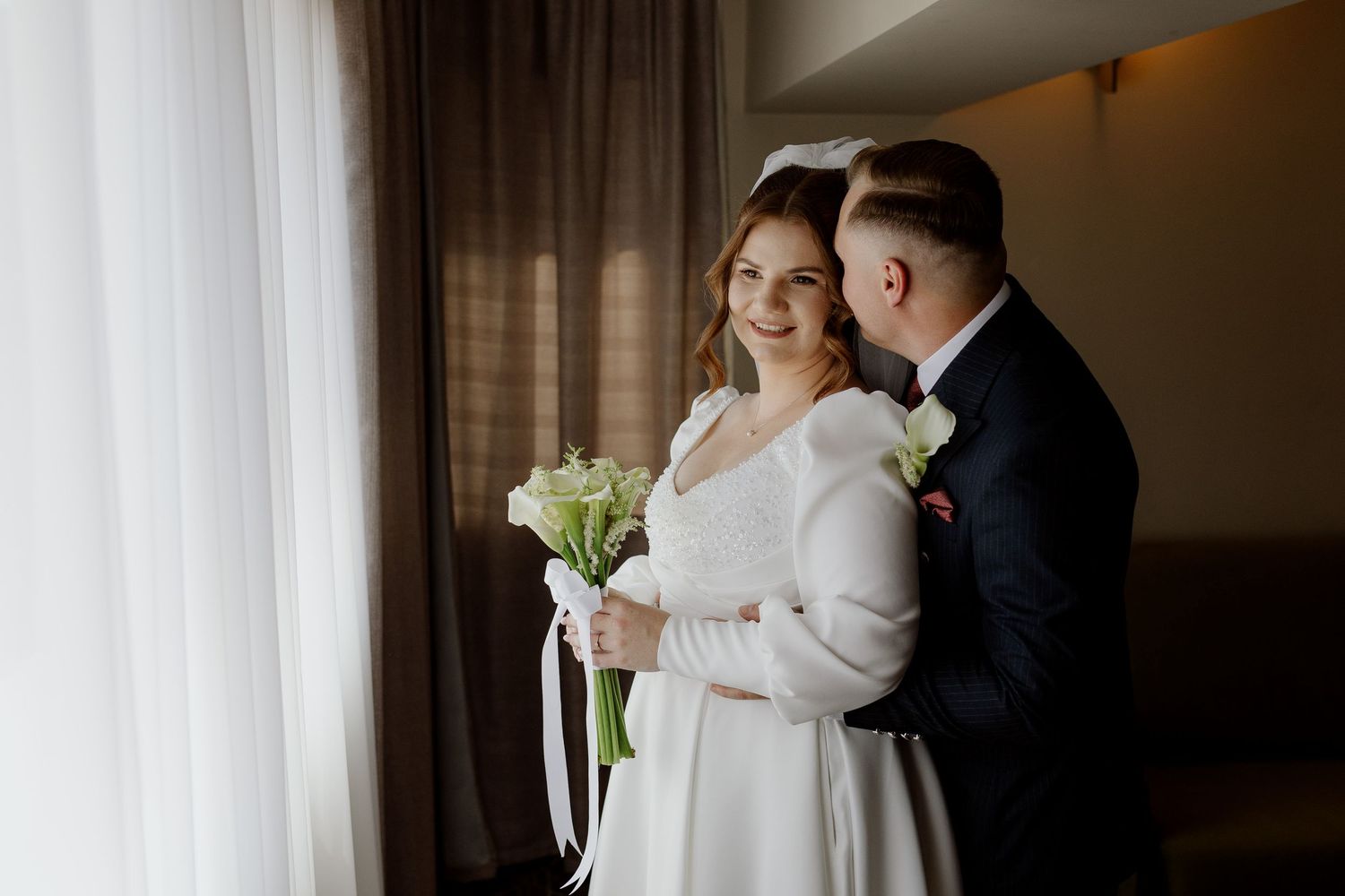 A bride and groom share an intimate moment in a softly lit room with white curtains.