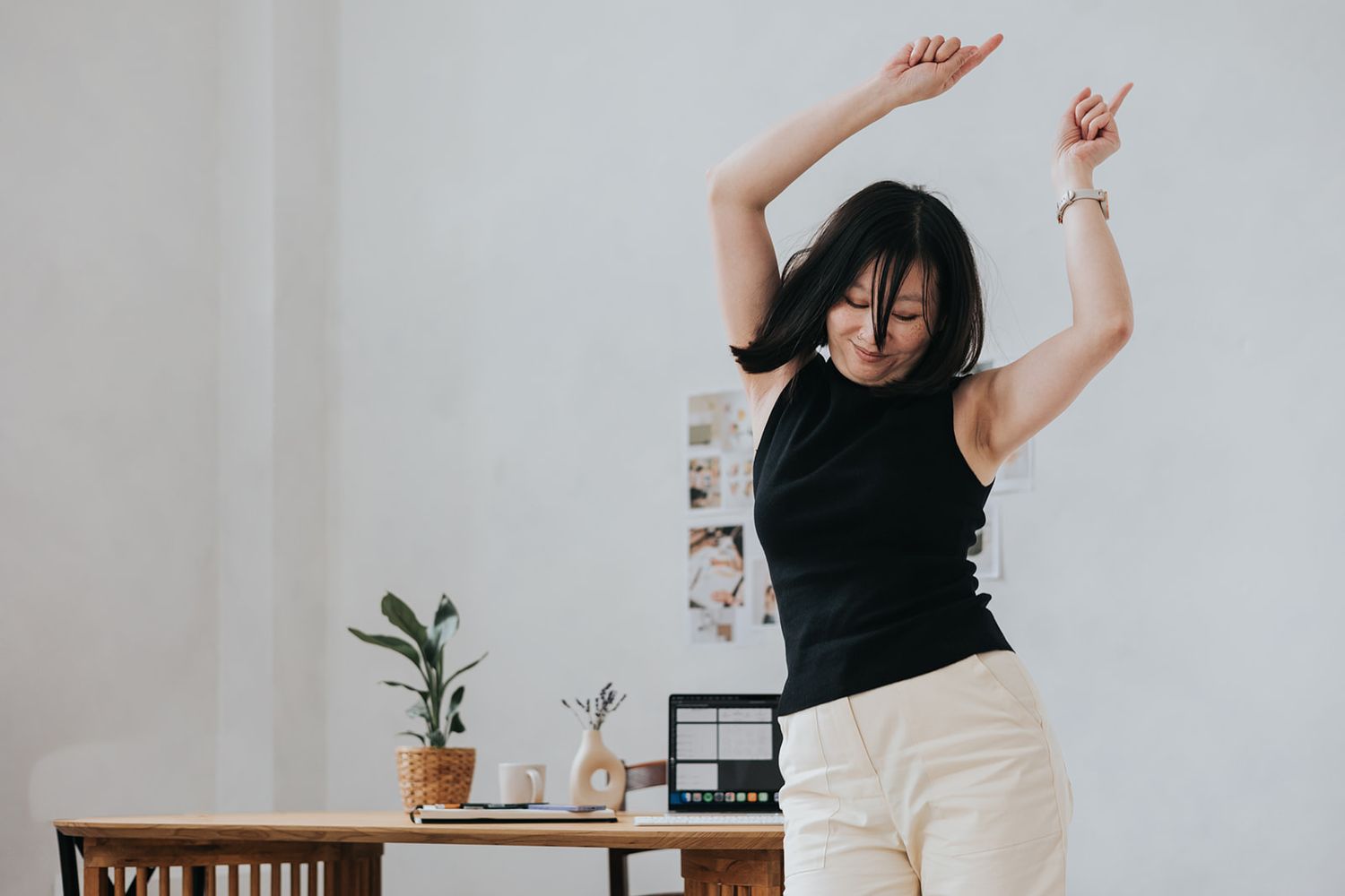 Woman in black sleeveless top and white pants dancing joyfully in a minimalist home office setting.