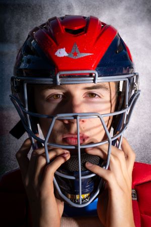 Close up of baseball catcher wearing red helmet with protective face mask against dramatic smoky background.