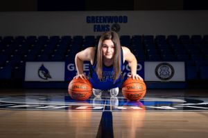 Basketball player in blue uniform poses with two basketballs on court with Greenwood Class S banner visible in background.