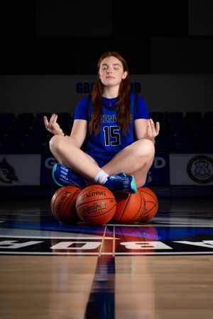A basketball player in blue jersey number 15 sits cross-legged on a court with basketballs in front.